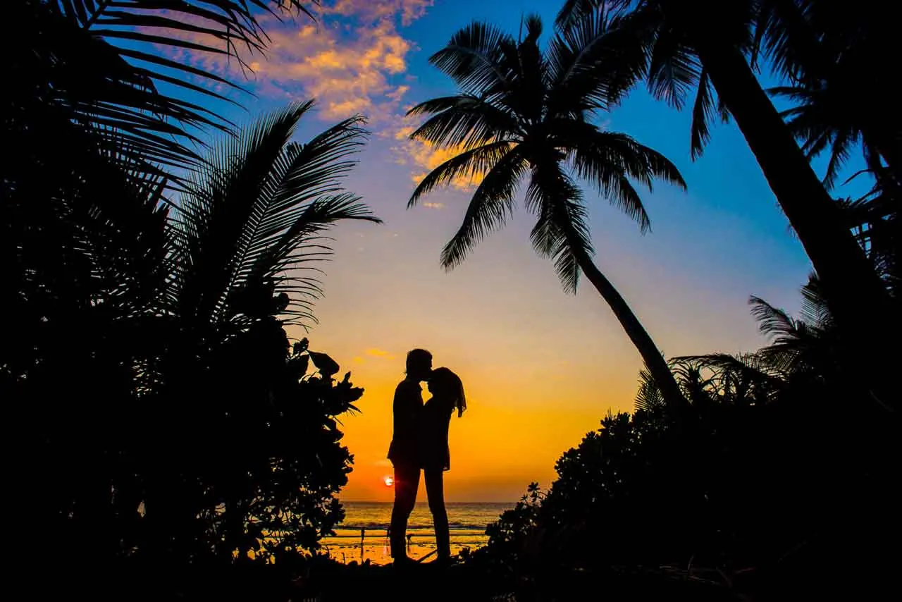 Silueta de una pareja que se besa en la playa al atardecer, entre palmeras